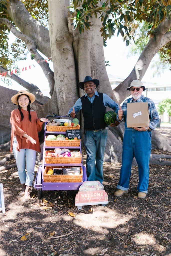 a-group-of-people-standing-near-the-cart-with-woven-baskets-8540985 Three diverse vendors at a farmers market showcasing fresh organic produce in a sunny outdoor setting.